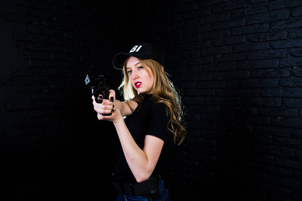 FBI female agent in cap and with gun at studio against dark brick wall. - Foto, imagen