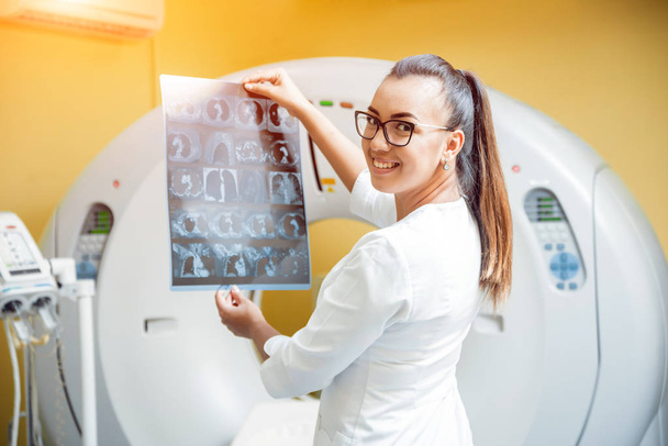 Female radiologist looking at x-ray in the room of computed tomography. Background - Фото, изображение