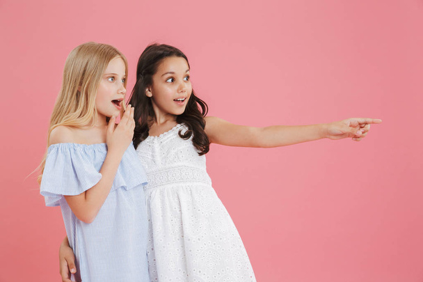 Image of two surprised and happy girls 8-10 years old wearing dresses looking aside and pointing finger at copyspace isolated over pink background - Foto, imagen