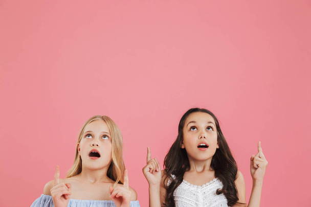 Photo of two surprised or shocked girls 8-10 years old in dresses looking upward with open mouth and pointing fingers at copyspace isolated over pink background - Фото, изображение
