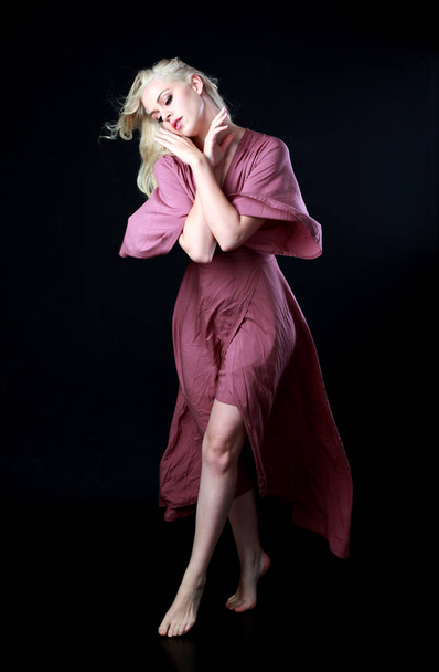full length portrait of blonde girl wearing long purple dress. standing pose. black studio background. - Photo, Image