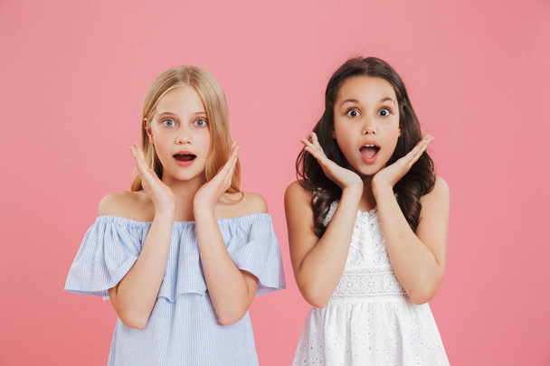 Photo of brunette and blonde princess girls wearing dresses expressing surprise or delight and raising hands at face with open mouth isolated over pink background - Photo, image