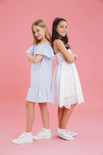 Full length photo of brunette and blonde girls wearing dresses smiling and looking at camera while standing back to back with arms folded isolated over pink background - Valokuva, kuva