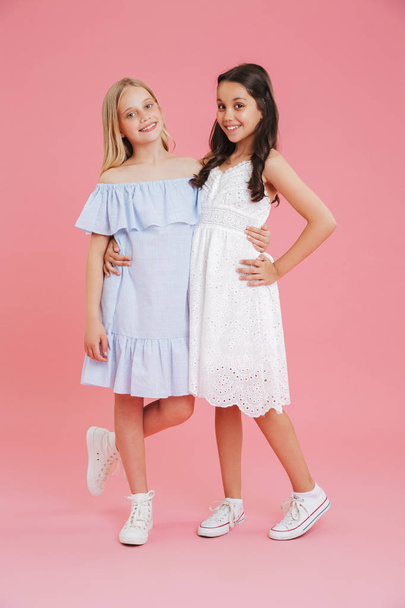 Full length photo of princess girls wearing dresses smiling and looking at camera while hugging together isolated over pink background - Valokuva, kuva