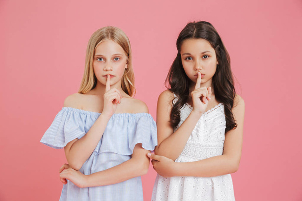 Photo of serious little girls 8-10 years old wearing dresses holding index finger on lips and asking to keep silence isolated over pink background - Foto, Bild