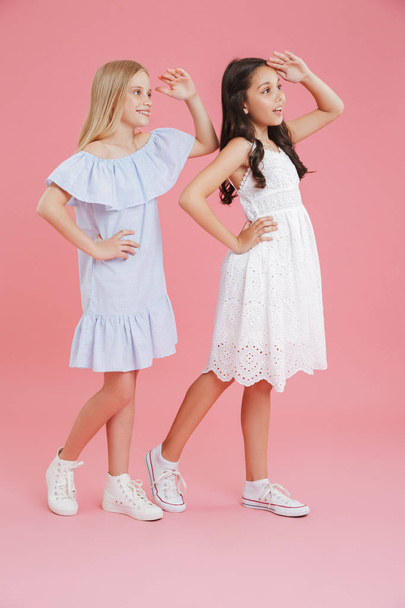 Full length photo of joyful girls wearing dresses smiling and looking far away with hands at their foreheads isolated over pink background - Фото, зображення