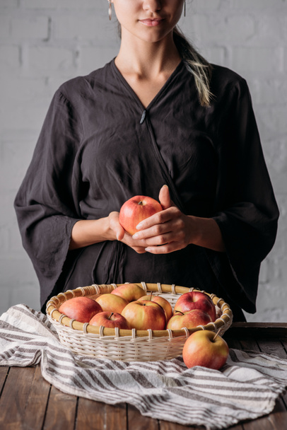 リネンの木製卓上にアップルと新鮮なりんごがいっぱい入ったかごを持つ女性の部分的なビュー - 写真・画像