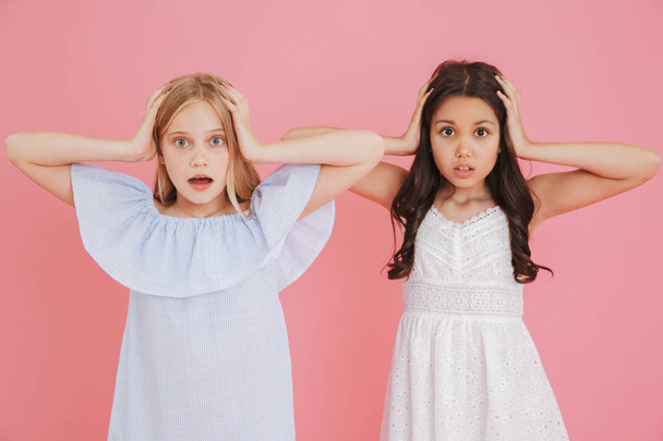 Photo of two shocked or scared girls 8-10 wearing dresses covering ears or grabbing heads with open mouth isolated over pink background - Foto, imagen