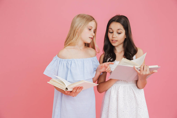 Picture of brunette and blonde smart girls wearing dresses holding and reading books together with interest isolated over pink background - Foto, Imagen
