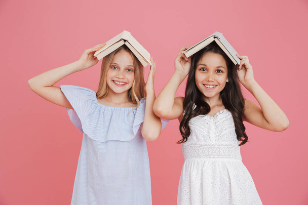 Picture of happy european girls wearing dresses holding books at their heads while smiling at camera isolated over pink background - Zdjęcie, obraz