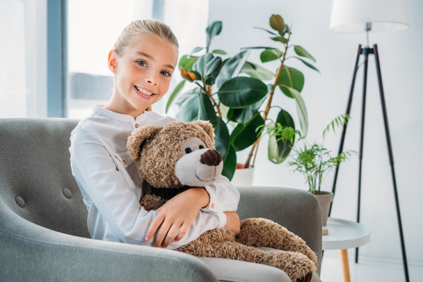 adorable little child embracing her teddy bear while sitting in armchair and looking at camera - Foto, Bild