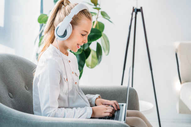side view of smiling little child in headphones using laptop while sitting in armchair - Photo, Image