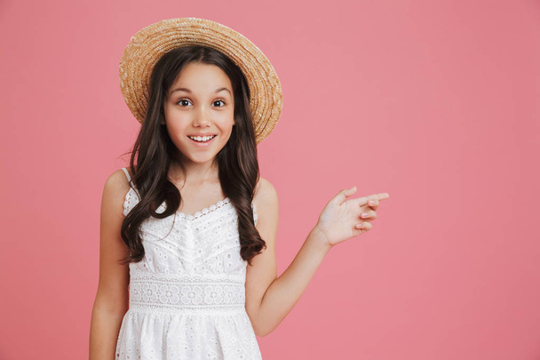 Portrait of brunette smiling girl 8-10 wearing white dress and straw hat looking at camera and pointing finger aside at copyspace isolated over pink background - Φωτογραφία, εικόνα