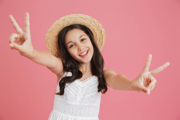 Excited european girl 8-10 wearing white dress and straw hat looking at camera while showing peace sign with both hands isolated over pink background - Photo, Image