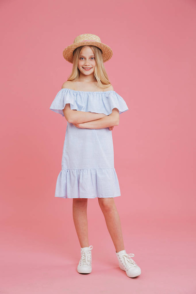 Full length portrait of beautiful blonde summer girl 8-10 wearing blue dress and straw hat posing at camera with arms crossed and smile isolated over pink background - Photo, Image