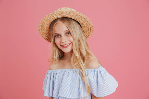 Image of lovely blond summer girl 8-10 wearing blue dress and straw hat smiling at camera with joyful glance isolated over pink background - Foto, imagen