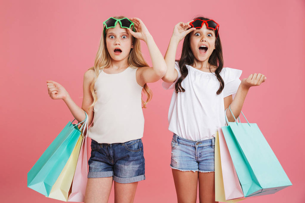 Foto van verbaasd brunette en blonde meisjes 8-10 in casual kleding op zoek onder de zonnebril terwijl kleurrijke boodschappentassen met aankopen geïsoleerd op roze achtergrond - Foto, afbeelding