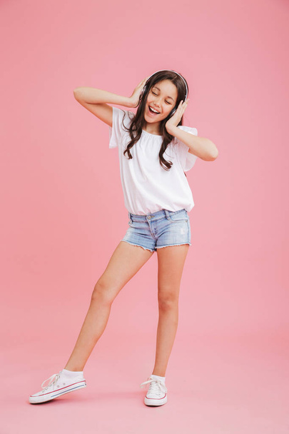 Full length portrait of cheerful girl 8-10 in casual clothing singing while listening to music via wireless headphones isolated over pink background - Photo, Image