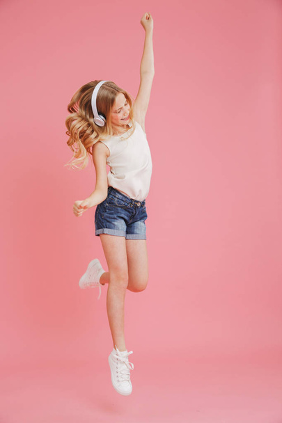 Full length image of blonde girl 8-10 in casual clothing dancing and listening to music with wireless headphones at head isolated over pink background - Fotó, kép