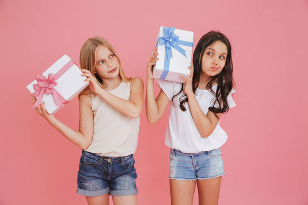 Two curious interested girls 8-10 in casual clothing holding and shaking gift boxes with colorful bows isolated over pink background - Photo, image