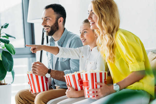 onnellinen nuori perhe katsomassa elokuvaa kotona ämpäreitä popcornia ja osoittaa televisioon
 - Valokuva, kuva