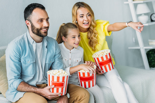 lächelnde junge Familie, die zu Hause mit Eimern Popcorn einen Film anschaut und auf den Fernseher zeigt - Foto, Bild
