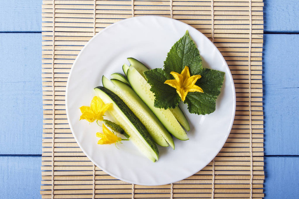 Sliceb cucumbers and leaves with yellow flowers on white plate. Top view on wooden table with bamboo mat. - Foto, imagen