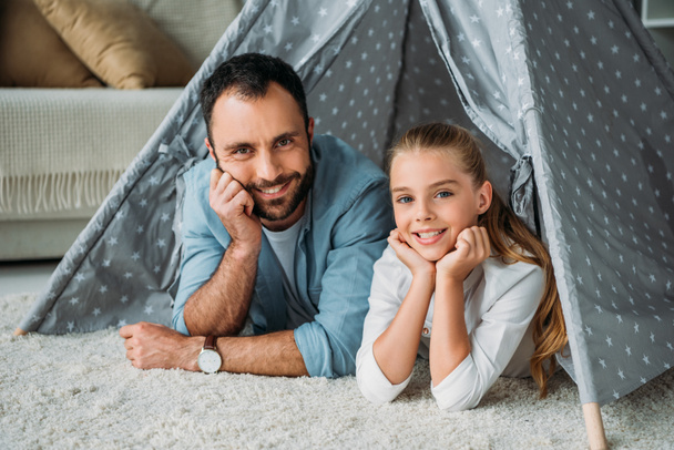 father and daughter lying on floor inside of teepee and looking at camera at home - Фото, изображение