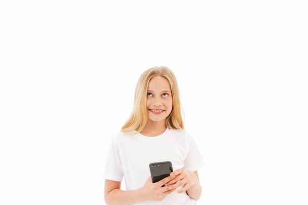 Happy pensive young blonde girl in casual clothes holding smartphone and looking away over white background - Photo, image