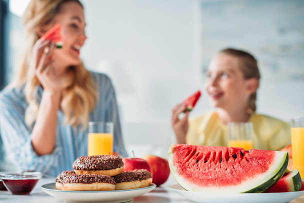 selective focus shot of mother and daughter having breakfast with donuts and watermelon on foreground at home - Foto, Imagen