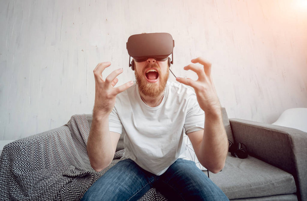 Young beardy man playing games with virtual reality goggles at home. VR technologies - Φωτογραφία, εικόνα