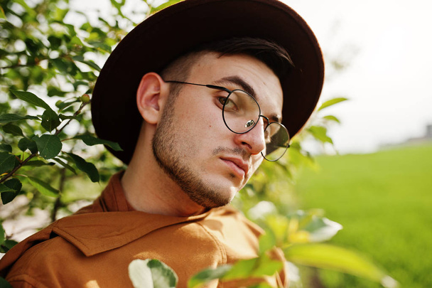 Stylish man in glasses, brown jacket and hat posed on green field. - Fotó, kép