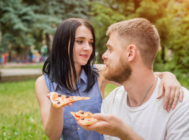 Νεαρό ζευγάρι τρώει πίτσα στο πάρκο. Μεταχειρίζονται κάθε άλλο και γελούν. - Φωτογραφία, εικόνα