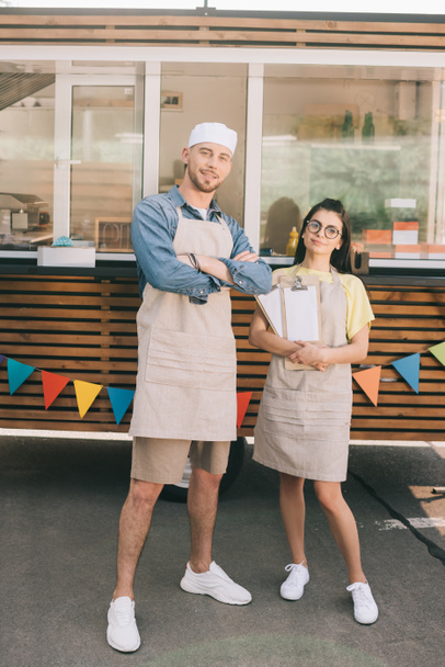 young small business owners in aprons standing with crossed arms and smiling at camera near food truck - Photo, Image