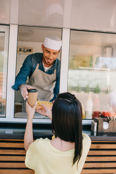 Улыбающийся молодой шеф-повар дает картофель фри и кофе, чтобы пойти к клиентке в фургоне с едой
 - Фото, изображение