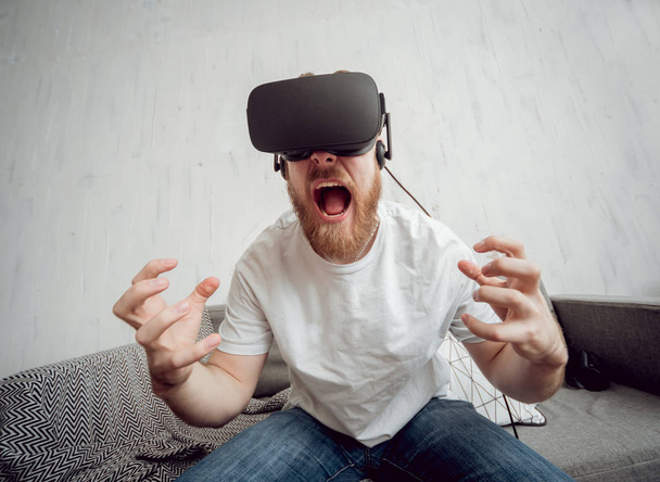 Young beardy man playing games with virtual reality goggles at home. VR technologies - Foto, afbeelding