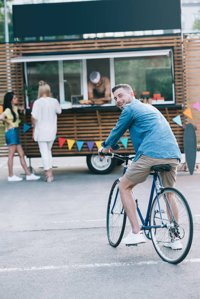 przystojny mężczyzna siedzi na rowerze w pobliżu food truck i patrząc na kamery - Zdjęcie, obraz