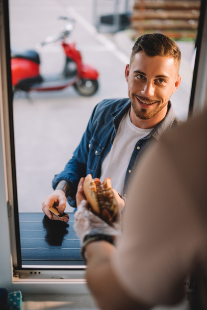 abgeschnittenes Bild des Chefs, der einem gutaussehenden Kunden in einem Foodtruck leckeren Hot Dog gibt  - Foto, Bild