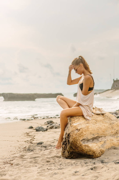 Woman natural beauty. Pretty girl sit on big stone. Young lady on exotic sand beach at sea coast enjoying sunset or ocean sunrise. Travel, surfer relax and active yoga meditating lifestyle concept. - 写真・画像