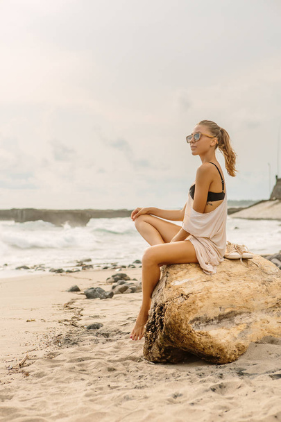 Woman natural beauty. Pretty girl sit on stone and look forward. Young lady on beach at sea coast enjoying sunset or ocean sunrise. Travel, surfer relax and active yoga meditating lifestyle concept. - 写真・画像