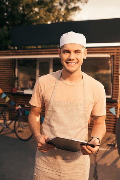 uśmiechający się przystojny szef kuchni w fartuch przytrzymanie schowka i patrząc na kamery w pobliżu food trucka - Zdjęcie, obraz