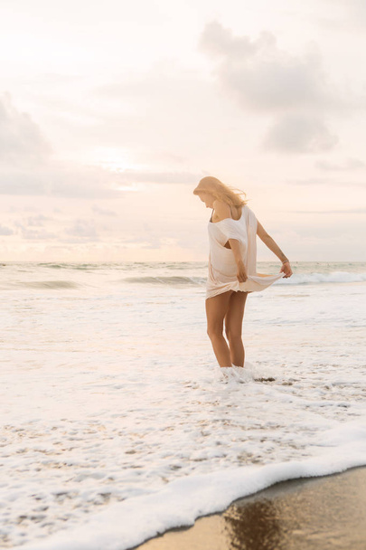 Hipster trendy girl hold her casual summer dress and walk in little waves on the coastline. Sporty lady on sea sand beach sunset or ocean sunrise. Travel, active, yoga lifestyle concept. - Photo, image