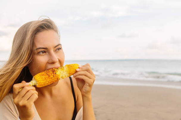 Healthy vegetarian hipster girl in summer casual outfit eat fresh grilled corn. Sexy lady on sea beach sunset or ocean sunrise. Travel, explore, active yoga and meditation lifestyle, dieting concept. - Foto, immagini