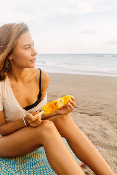 Healthy diet. Vegetarian hipster girl eat fresh organic grilled corn. Sexy lady on sea beach sunset or ocean sunrise. Travel, explore, active, yoga, vegan and meditation lifestyle, dieting concept. - Photo, image