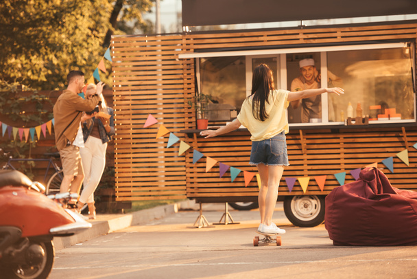πίσω όψη του κορίτσι στέκεται για skateboard κοντά φορτηγό τροφίμων - Φωτογραφία, εικόνα