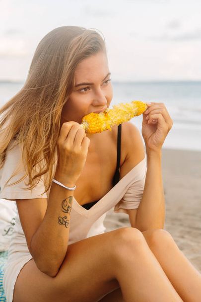 Healthy diet. Vegetarian hipster woman in summer outfit eat fresh organic grilled corn. Sexy lady on sea beach sunset or ocean sunrise. Travel, explore, active yoga and meditation lifestyle concept. - Foto, Imagen