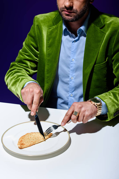 abgeschnittene Aufnahme eines Mannes in stilvoller grüner Samtjacke beim Schneiden von Fleischgebäck auf einem Teller mit blauem Hintergrund dahinter - Foto, Bild
