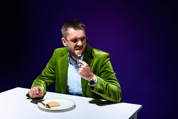 portrait of man in stylish green velvet jacket eating meat pastry on plate at table with blue background behind - Photo, Image