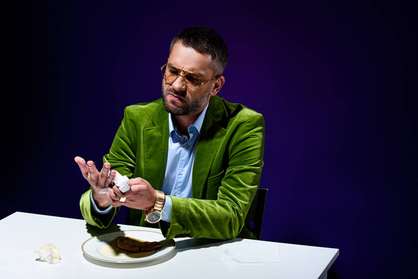 man in stylish green velvet jacket drying hands with napkin at table with meat pastry on plate with blue background behind - Foto, Imagen