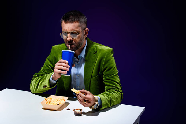fashionable man in velvet jacket drinking soda drink while sitting at table with junk food with blue backdrop - Photo, Image